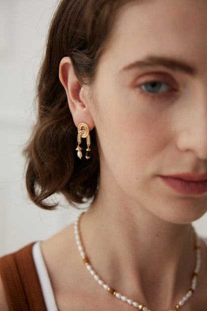 Close-up of a woman with short brown hair wearing Siren Mode's Vintage Gold Tiger's Eye and Pearl Drop Earrings, featuring dangling white and black beads. She also wears a beaded necklace with alternating gold and white beads. She is dressed in a brown tank top layered over a white top, accentuating her natural pearl earrings.