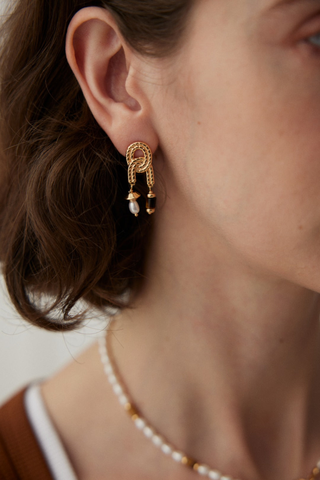 Close-up of a person wearing Siren Mode's Vintage Gold Tiger's Eye and Pearl Drop Earrings, which are shaped like two connected loops, adorned with small black beads and a dangling natural pearl. The person's short brown hair partially obscures the ear, and a delicate beaded necklace is visible around their neck.