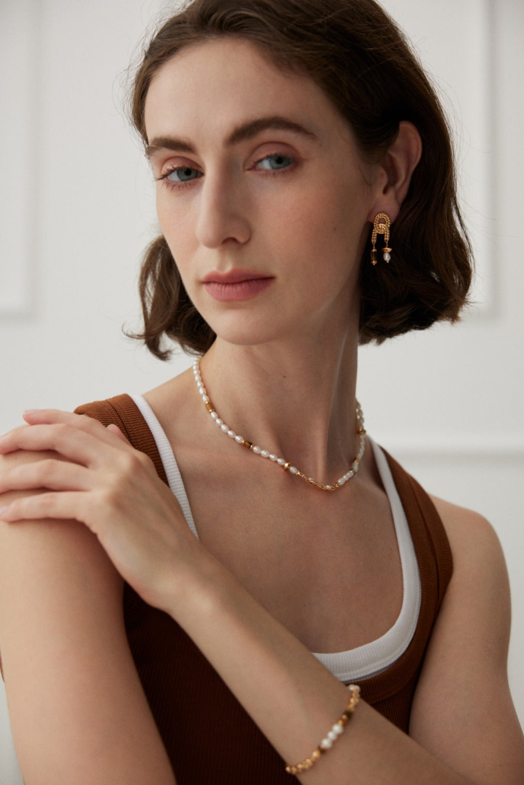 A person with short, wavy hair is wearing a brown and white tank top, featuring 925 silver jewelry including a necklace, bracelet, and Siren Mode's Vintage Gold Tiger's Eye and Pearl Drop Earrings. The background is white, and their expression is calm and composed.