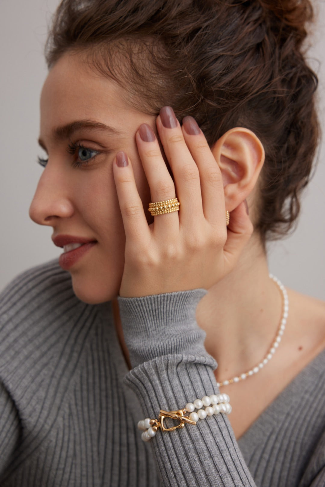 A person with brown hair pulled back is shown in profile, resting their hand on their face. They are wearing a Siren Mode Vintage Gold and White Gold 925 Silver Adjustable Beaded Ring, a pearl bracelet with a gold clasp, and a gray ribbed sweater. Their nails are painted in a muted mauve color.