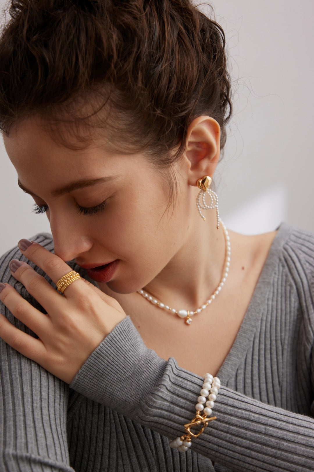 A woman with brown hair tied back is wearing a grey ribbed sweater and various pieces of jewelry, including a pair of gold earrings, a pearl necklace, a gold ring, and a bracelet with pearls. She looks down with her hand gently touching her face, showcasing the Vintage Gold and White Gold 925 Silver Adjustable Beaded Rings by Siren Mode.