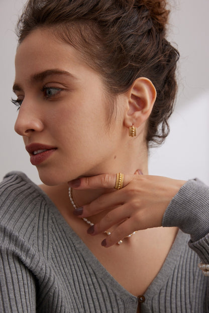 A woman with her hair in an updo is looking to the side. She wears gold hoop earrings, a Vintage Gold and White Gold 925 Silver Adjustable Beaded Ring from Siren Mode, and a gray ribbed sweater. Her hand, which rests near her chin, shows neatly manicured nails. The background is softly lit and neutral-colored.