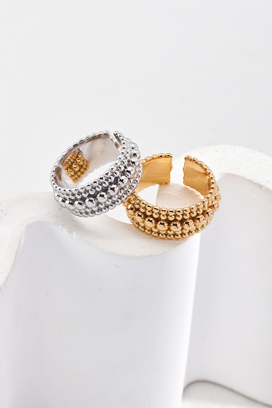 Two intricately designed adjustable rings from Siren Mode's Vintage Gold and White Gold 925 Silver Adjustable Beaded Rings collection are displayed against a white background. One ring, showcasing the elegance of vintage gold beaded rings, and the other in silver, feature multiple rows of small, textured beads along their bands. The rings are shown resting on a white, wavy surface.