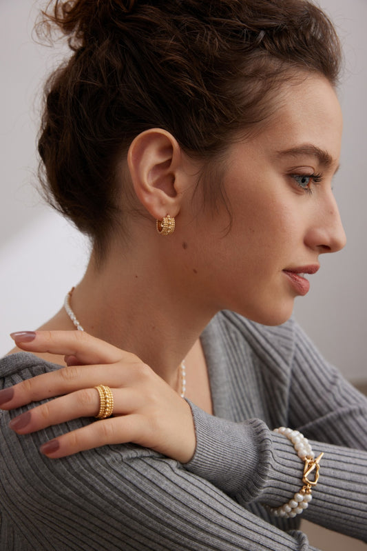 A woman with wavy brown hair is shown in profile, wearing a gray top. She has Siren Mode's Vintage Gold and White Gold 925 Silver Beaded Hoop Earrings, a gold ring, and a pearl bracelet with a gold clasp on her right hand, which rests on her left shoulder. She appears thoughtful, looking slightly downward.