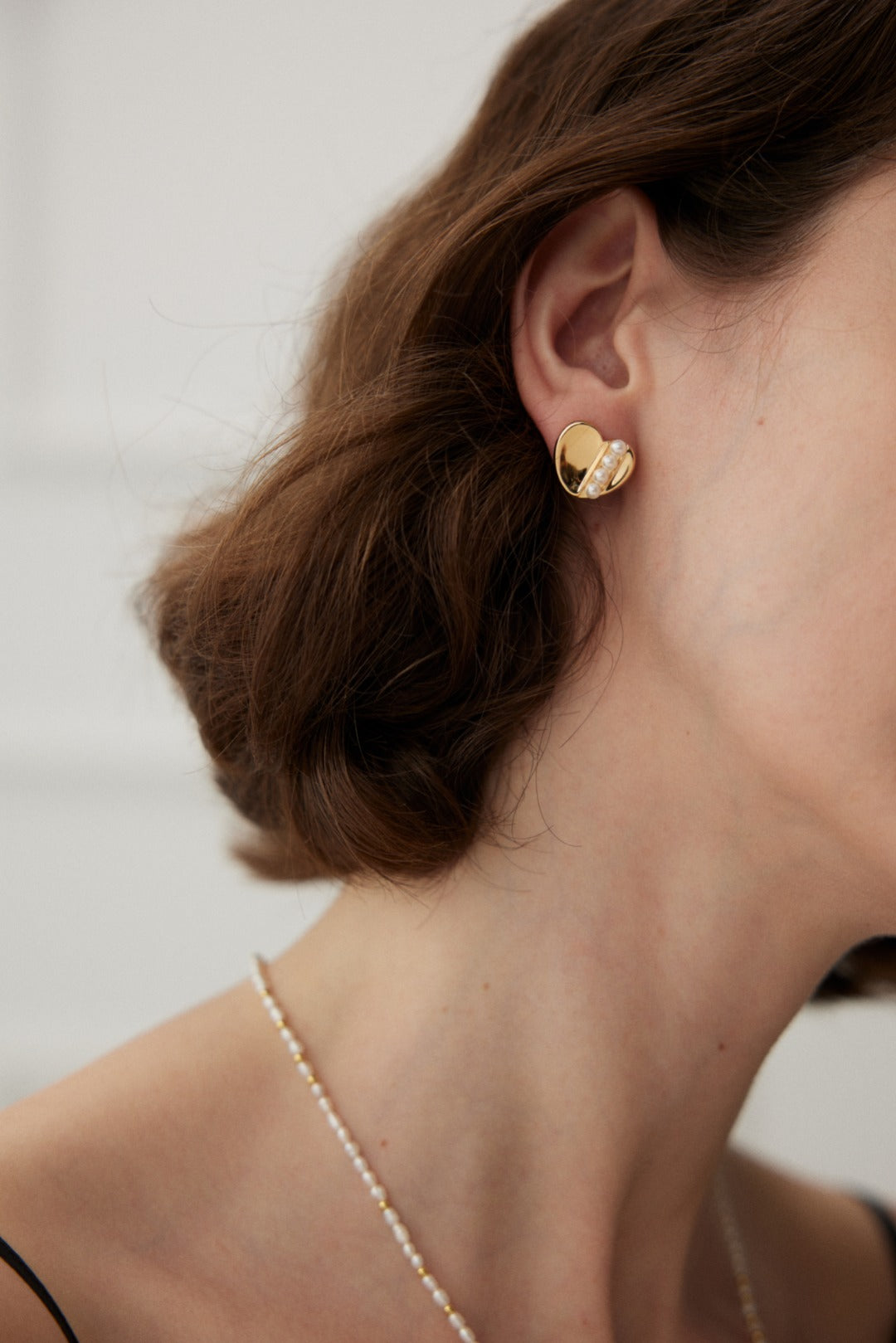 Close-up of a person wearing Siren Mode's Vintage Gold and White Gold 925 Silver Heart Pearl Stud Earrings. The person has wavy brown hair styled in a bob and is wearing a thin necklace adorned with small beads. The background is blurred and white.