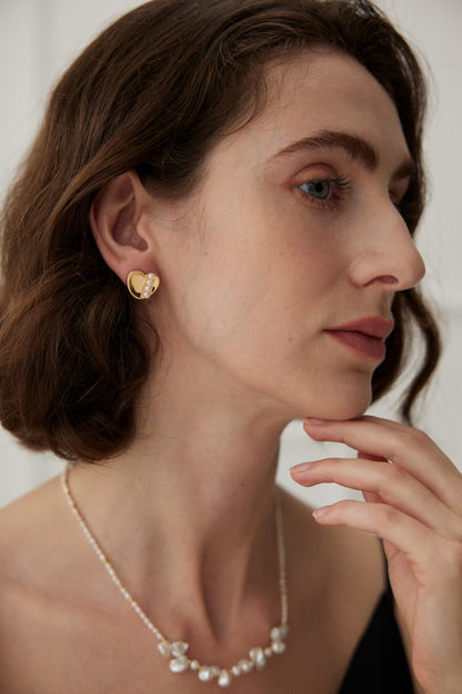 A woman with shoulder-length brown hair is shown in a close-up profile view. She wears a pair of Siren Mode Vintage Gold and White Gold 925 Silver Heart Pearl Stud Earrings and a delicate pearl necklace. Her hand is gently touching her chin, and she is wearing natural makeup. The background is a soft, light color.