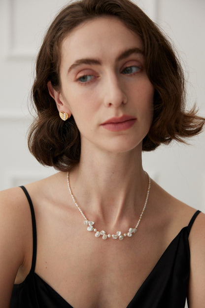 A woman with short, wavy brown hair wears a black spaghetti strap top, Siren Mode Vintage Gold and White Gold 925 Silver Heart Pearl Stud Earrings, and a delicate beaded necklace. She has a neutral expression and is looking slightly to the side. The background is a softly-lit white wall with panel details.