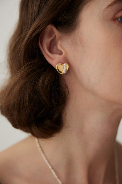 Close-up of a person wearing Siren Mode's Vintage Gold and White Gold 925 Silver Heart Pearl Stud Earrings. The person has short, wavy brown hair and is also wearing a delicate necklace with small, light-colored beads. The background is out of focus, emphasizing the jewelry.