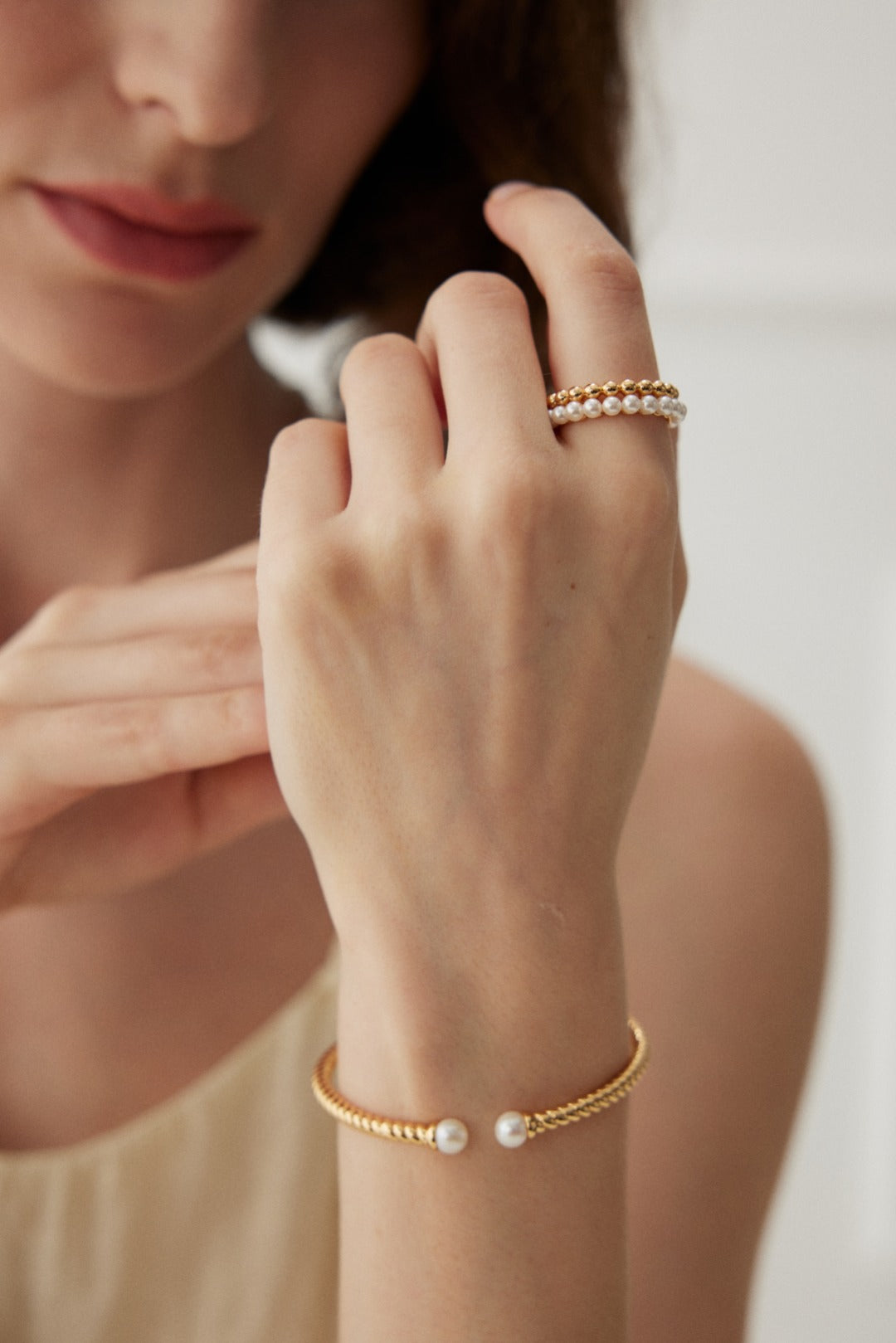 A person with light skin is wearing a gold bracelet adorned with pearls on their wrist and a Siren Mode Vintage Gold and White Gold 925 Silver Pearl Beaded Ring on their finger. The background is out of focus, emphasizing the jewelry. The person is lightly touching their chin with their other hand.