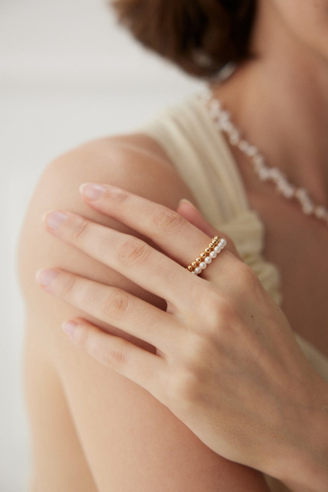 Close-up of a person with light skin tone, wearing a Siren Mode Vintage Gold and White Gold 925 Silver Pearl Beaded Ring. The person is gently touching their shoulder with their hand, showcasing the ring. They are also wearing a pearl beaded necklace. The background is out of focus, emphasizing the jewelry.