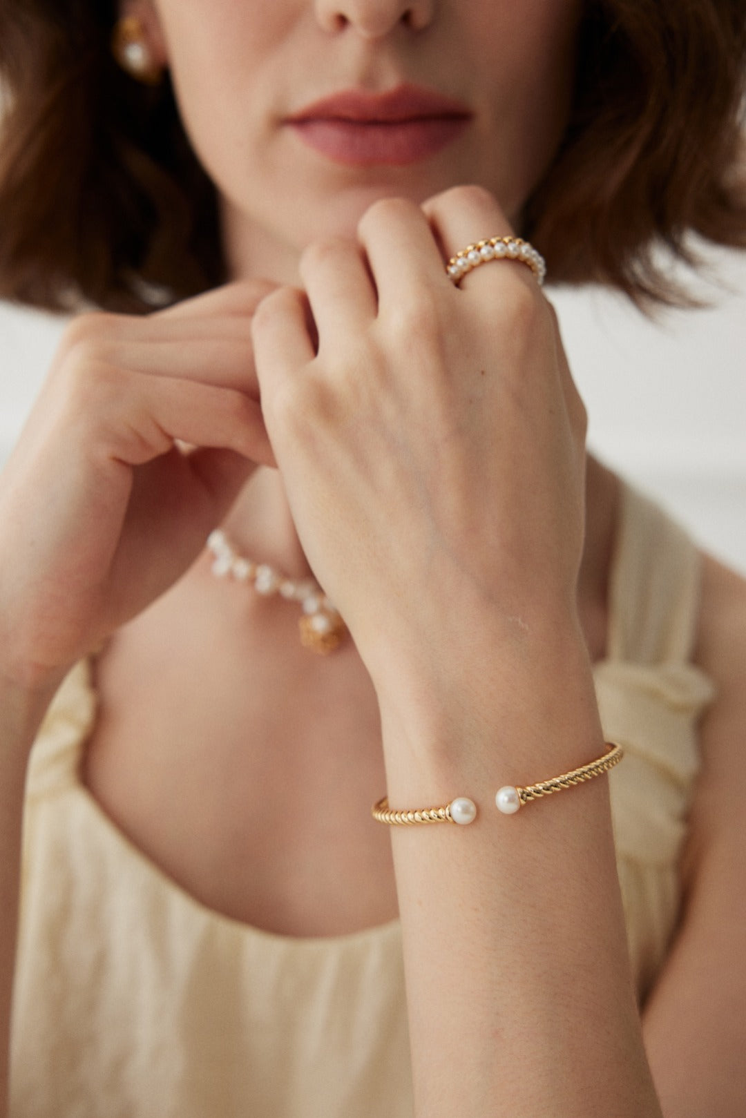 A close-up of a woman with light brown hair wearing a gold bracelet adorned with pearls on her wrist. She also wears a pearl necklace and two rings—one is the Siren Mode Vintage Gold and White Gold 925 Silver Pearl Beaded Ring, while the other features an elegant gold design. She is dressed in a light cream-colored top.