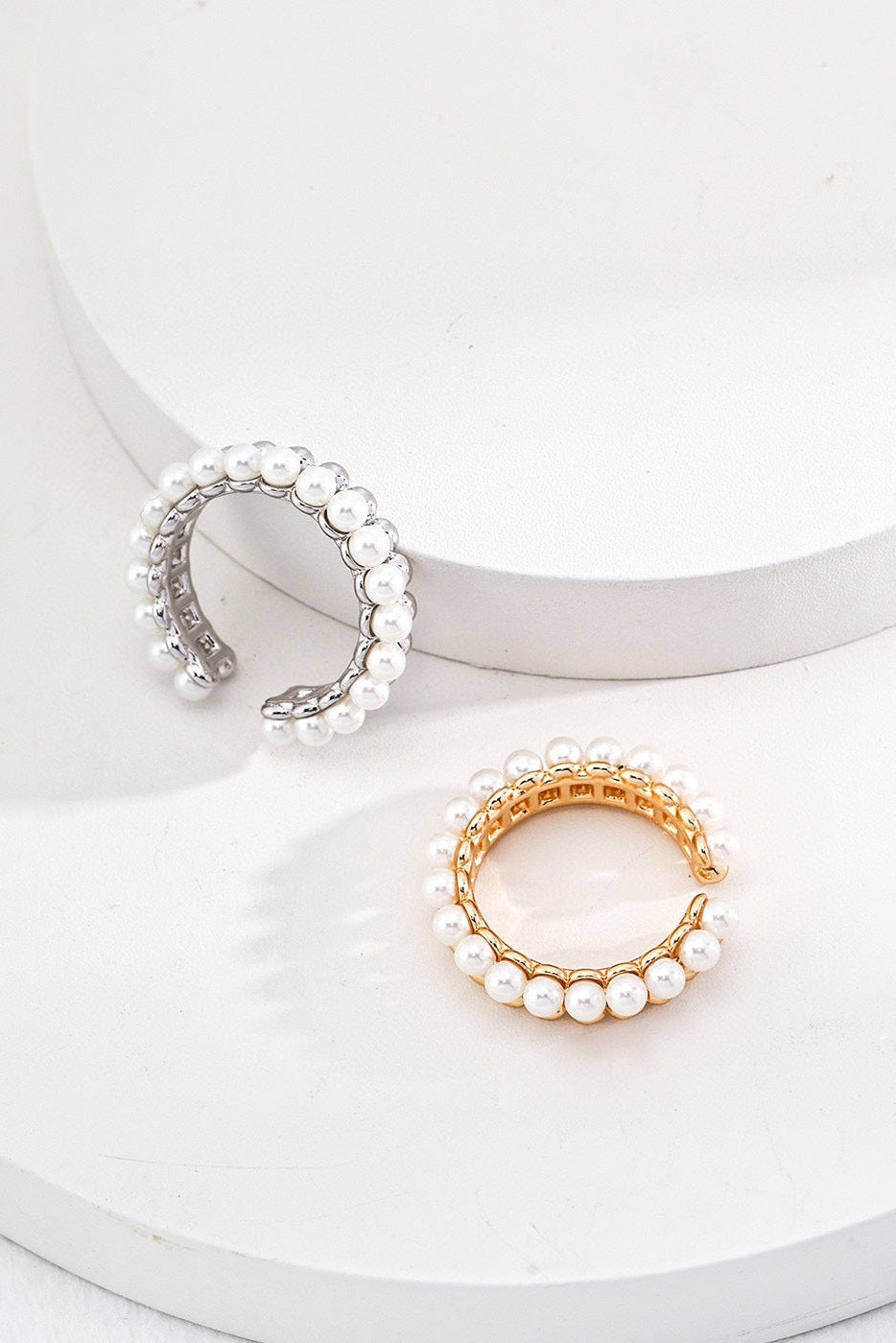 Displayed on a white, round podium are the exquisite Vintage Gold and White Gold 925 Silver Pearl Beaded Rings from Siren Mode. One ring is crafted in gold and the other in silver, both beautifully embellished with delicate white pearls along the outer edge. The gold ring, reminiscent of a vintage design, gracefully sits atop the podium while its silver counterpart rests elegantly on a lower platform.