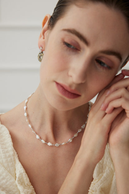 A woman with fair skin and slicked-back hair wears a beaded necklace and a decorative earring. She is dressed in a cream-colored textured top, gently holding one hand to her ear, looking down with a serene expression. Her exquisite White Gold 925 Silver Baroque Pearl Necklace from Siren Mode complements her elegant look.