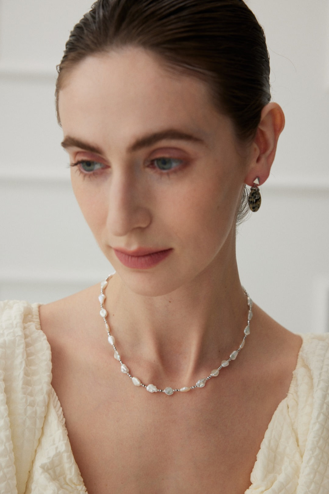 A woman with light skin and brown hair, styled in a pulled-back look, wears the Siren Mode White Gold 925 Silver Baroque Pearl Necklace along with a single stone earring. She is dressed in a textured off-white top, gazing downward with a serene expression against a softly blurred and bright background.