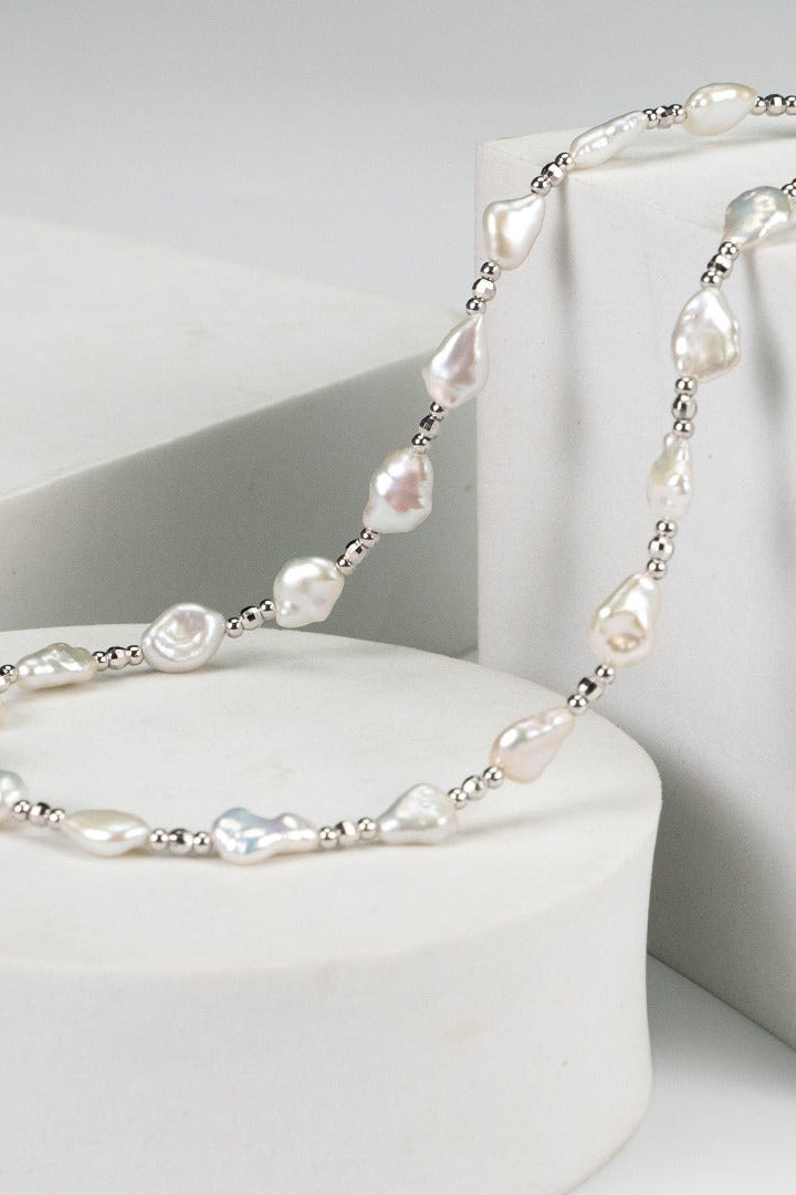 The Siren Mode White Gold 925 Silver Baroque Pearl Necklace, featuring unevenly shaped freshwater pearls, is elegantly displayed on white geometric pedestals. The baroque pearls cluster together, interspersed with small metallic beads, creating a harmonious blend of natural and crafted elements.