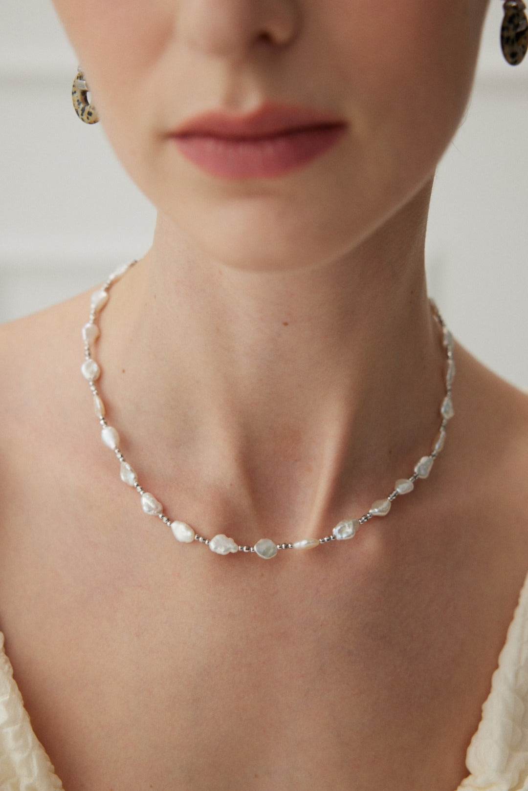 A close-up shot beautifully captures a woman's neck and lower face adorned with the Siren Mode White Gold 925 Silver Baroque Pearl Necklace. The necklace, featuring delicate baroque pearls and translucent beads, complements her fair complexion and cream-colored top against a softly blurred background.