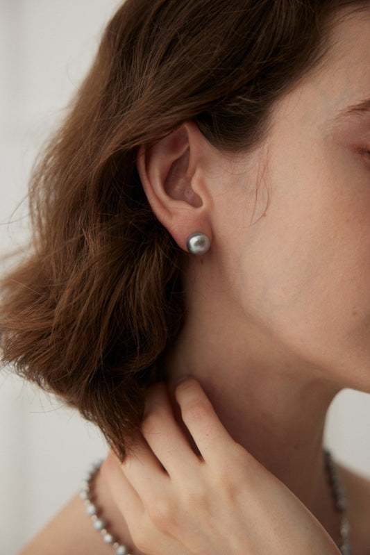 Close-up of a person wearing White Gold 925 Silver Natural Pearl Stud Earrings by Siren Mode. The person has short, wavy brown hair and holds their neck with one hand. The background is softly blurred, drawing attention to the earrings and the person's hair texture, highlighting the elegance of Siren Mode's 925 silver jewelry.