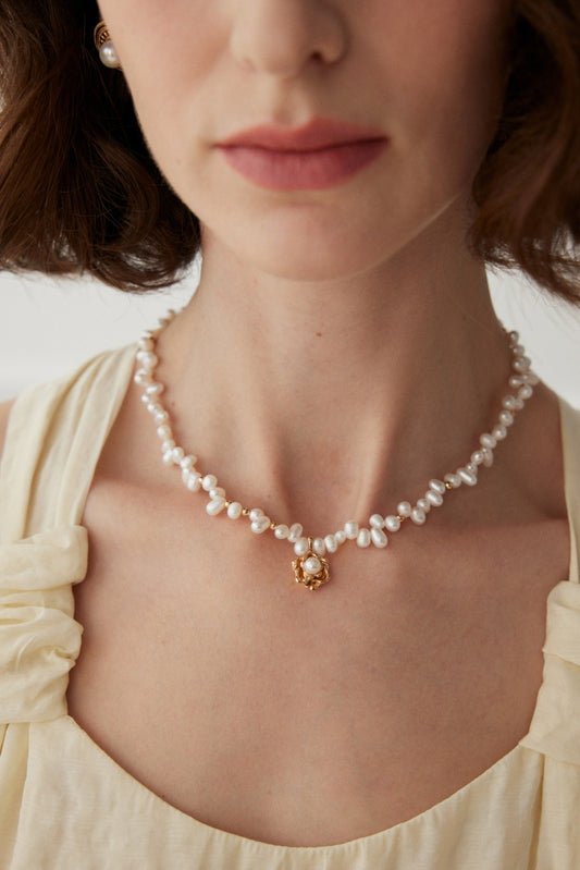 A close-up of a woman wearing a cream-colored top and the Elegant Vintage Gold Camellia Pearl Necklace from Siren Mode, featuring natural pearls and a 925 silver pendant with a centered pearl. The image focuses on her neck and collarbone, with her chin and rosy lips slightly visible at the top.